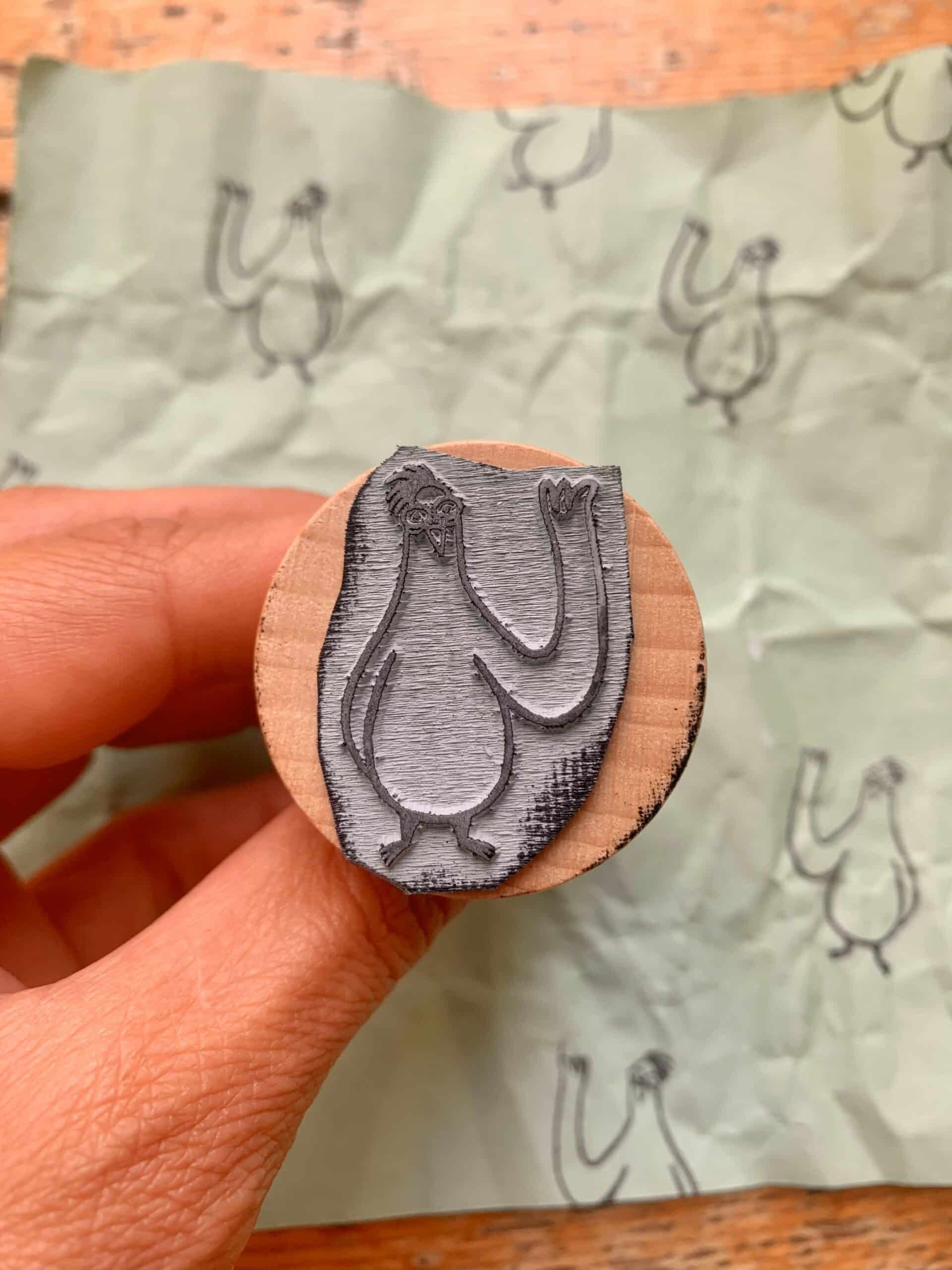 A person holding a wooden stamp of my one Comic „ A Pigeon’s Tale“ featuring a stylized pigeon design, with faint pigeon sketches in the background on crumpled green paper.